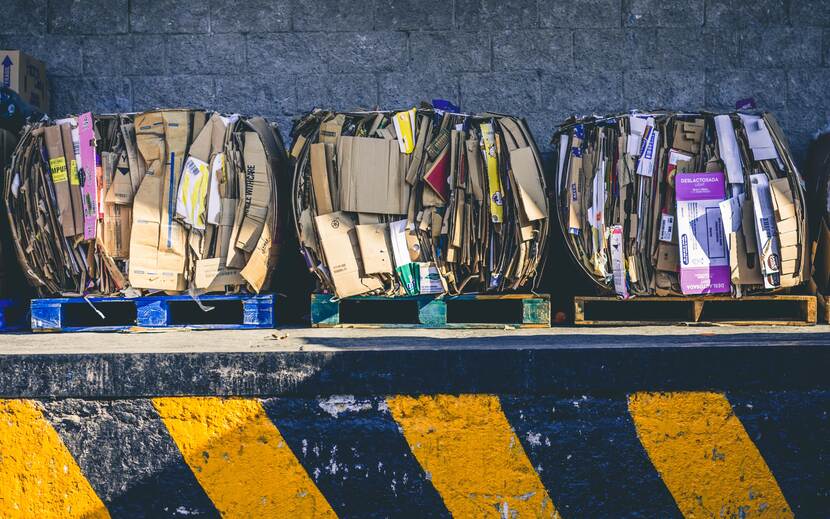 Illustratieve foto van drie balen samengeperst oud papier dat op pallets staat te wachten op transport. De drie pallets staan op een laaedplatform voor vrachtwagens, op de voorgrond zijn de geelzwarte waarschuwingsstrepen van de stootmuur zichtbaar.
