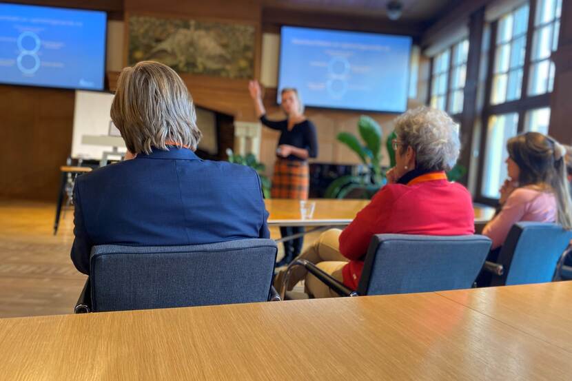 illustratieve afbeelding van het mini-symposium. Twee deelnemers op de rug gezien luisteren naar het verhaal van Marleen Hermans, die onscherp op de achtergrond van de foto te zien is.