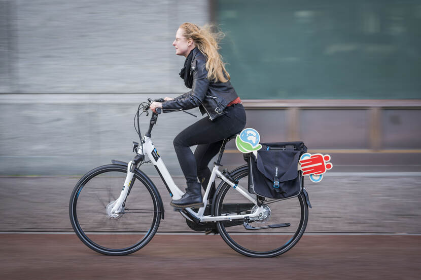 Anna van Mierlo fietst langs de glazen gevel van het ministerie van Buitenlandse Zaken in Den Haag. In haar fietstas heeft ze enkele voorwerpen die symbool staan voor het programma Denk Doe Duurzaam.