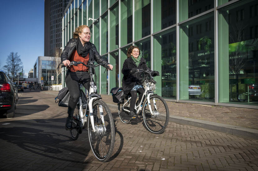 Anna van Mierlo (links) en Veronique Ruiz van Haperen (rechts) fietsen langs de glazen gevel van het ministerie van Buitenlandse Zaken in Den Haag.