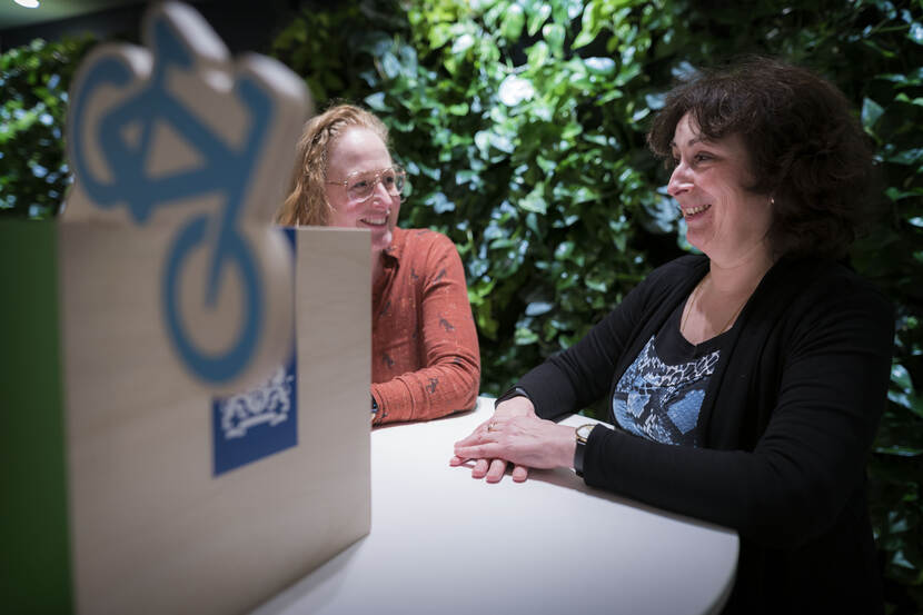 Anna van Mierlo (links) en Veronique Ruiz van Haperen in gesprek met elkaar aan een witte tafel. Op de achtergrond groene planten, links van beide dames een bak met daarin voorwerpen die symbool staan voor het programma Denk Doe Duurzaam.